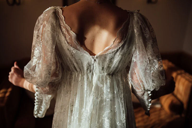 Dos d'une mariée portant une robe en dentelle blanche vaporeuse, éclairée par une lumière naturelle et douce.