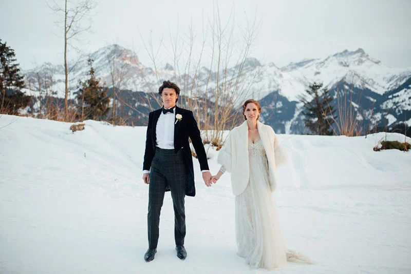 Couple marié main dans la main dans un paysage enneigé avec des montagnes en arrière-plan.