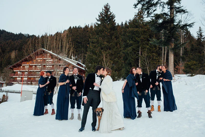 Mariés s'embrassant dans la neige entourés de leurs témoins en tenues traditionnelles bleues et noires devant un chalet.