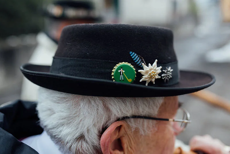 Gros plan sur un homme âgé portant un chapeau traditionnel noir orné de badges et d'une fleur blanche.