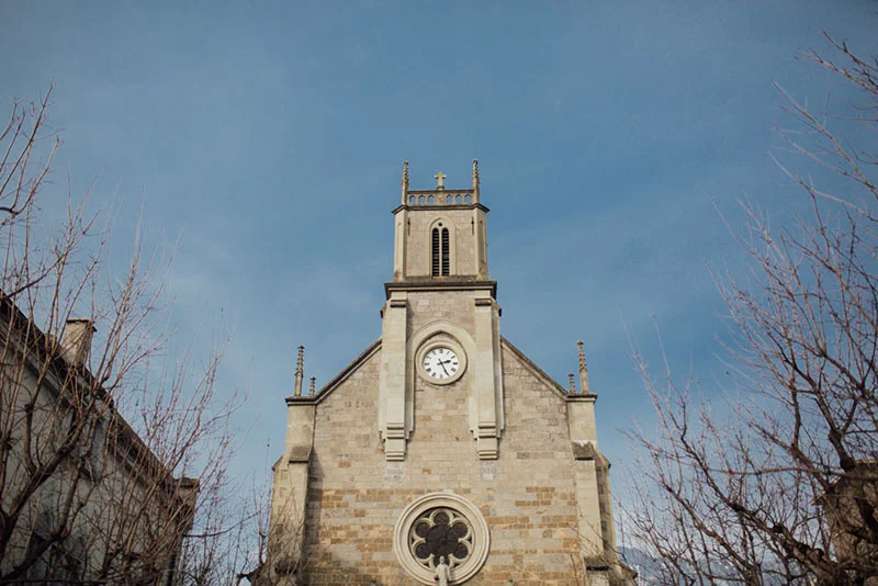 Façade en pierre d'une église ancienne sous un ciel bleu clair, encadrée par des branches d'arbres sans feuilles.