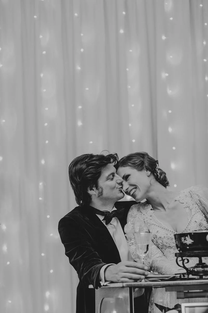 Couple de mariés complices et souriants, en noir et blanc, devant un rideau lumineux.