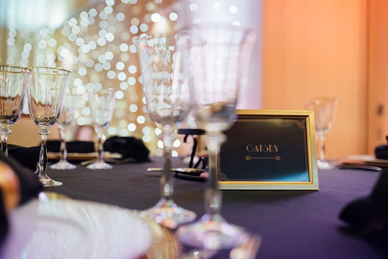 Table de mariage élégante avec verres en cristal, nappe sombre et marque-place Gatsby sur fond de lumières scintillantes.
