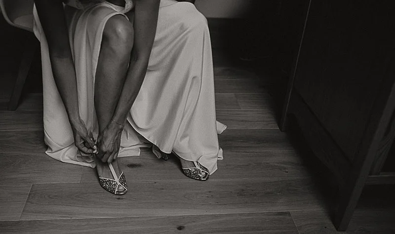 Une mariée en robe blanche ajuste ses chaussures scintillantes, vue en noir et blanc dans une ambiance intimiste.