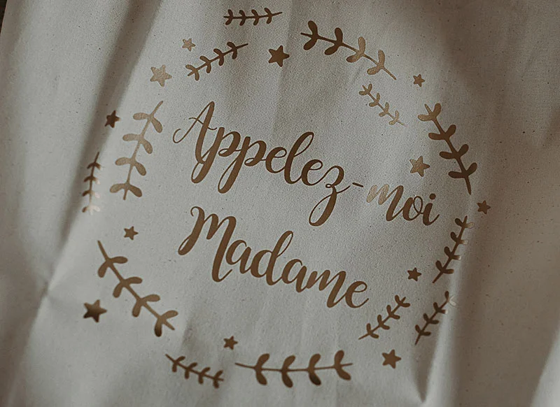 Texte doré « Appelez-moi Madame » entouré d'une couronne végétale sur un tissu blanc cassé.