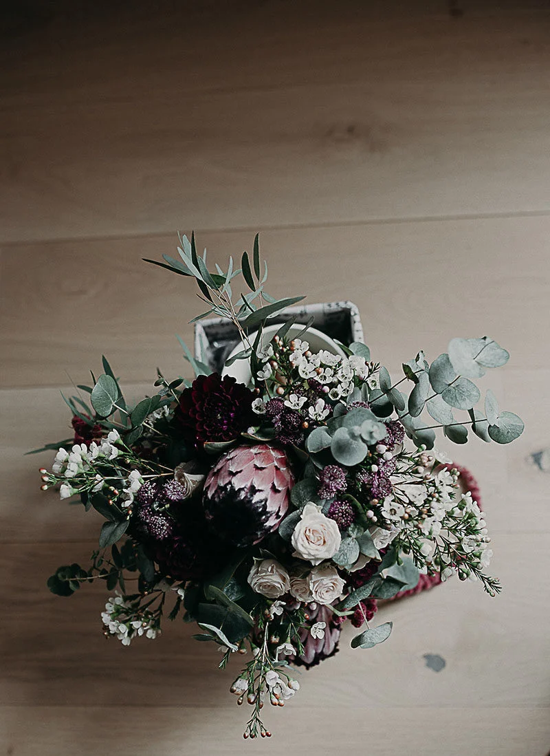 Bouquet de mariée aux tons bordeaux et blanc avec un artichaut, posé sur un sol en bois clair.