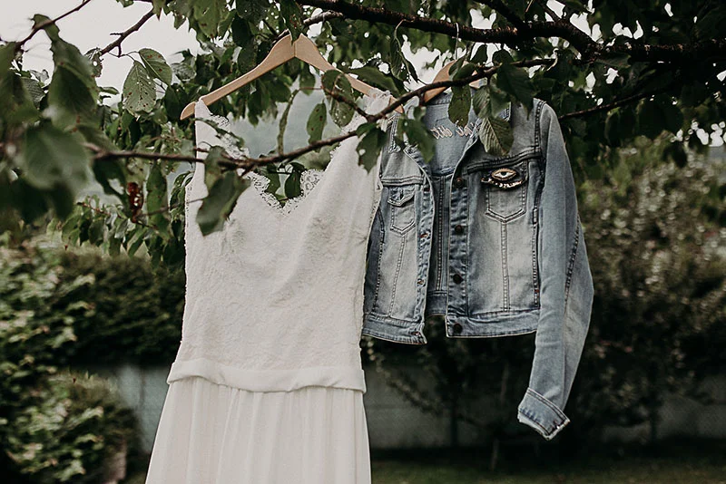Robe de mariée blanche et veste en jean suspendues à la branche d'un arbre dans un jardin verdoyant.