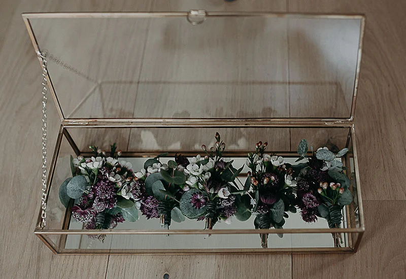 Boutonnières aux tons blancs et pourpres disposées dans une boîte en verre et métal doré sur un parquet clair.