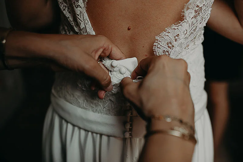 Gros plan sur des mains boutonnant le dos d'une robe de mariée en dentelle blanche.