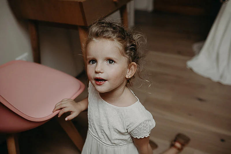 Petite fille aux cheveux bouclés portant une robe blanche, regardant avec curiosité dans un intérieur lumineux.