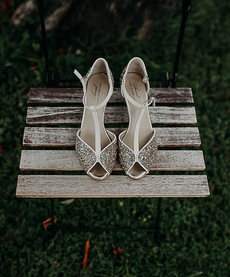 Paire de sandales à talons scintillantes et argentées posées sur un petit banc en bois rustique.