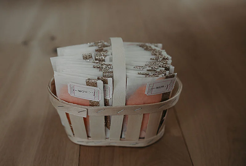 Petits cadeaux d'invités blancs et dorés disposés dans un panier en osier blanc sur un parquet en bois.