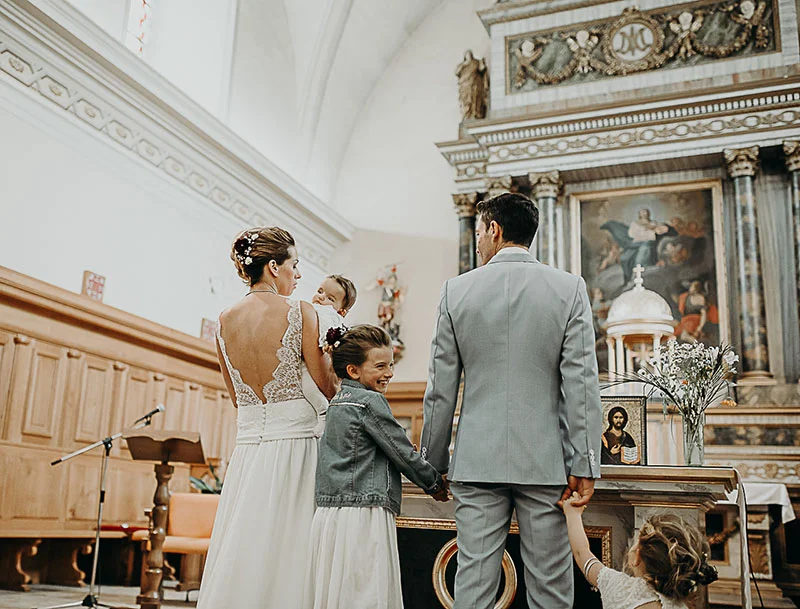 Mariés et enfants de dos face à l'autel d'une église lumineuse, ambiance solennelle et tendre.