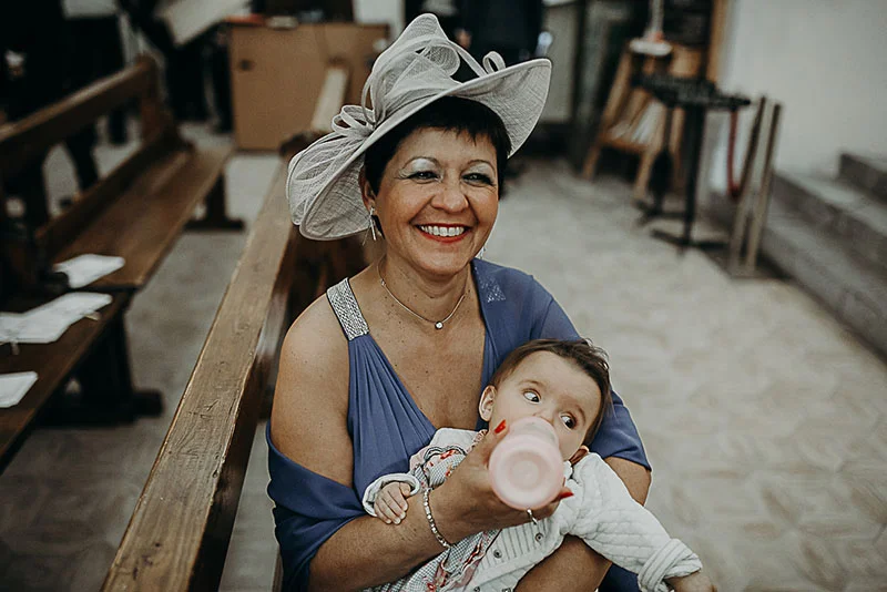 Une femme souriante portant un chapeau blanc et une robe bleue tient un bébé qui boit son biberon.