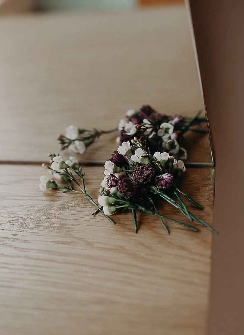 Boutonnières de fleurs blanches et violettes posées sur une table en bois clair.