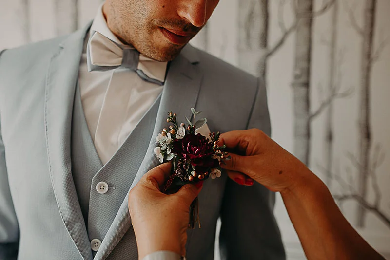 Une main ajuste une boutonnière aux fleurs bordeaux sur le costume bleu clair d'un marié.