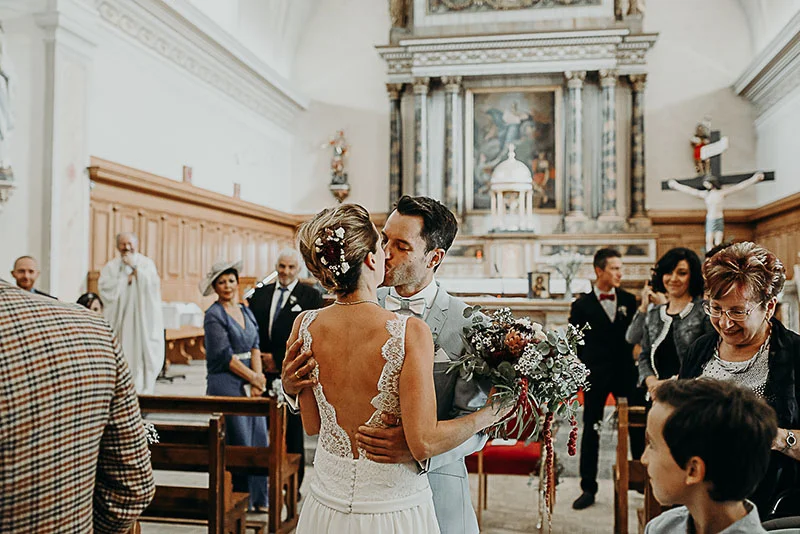 Un couple de mariés s'embrasse dans une église entouré de leurs proches dans une ambiance solennelle et chaleureuse.