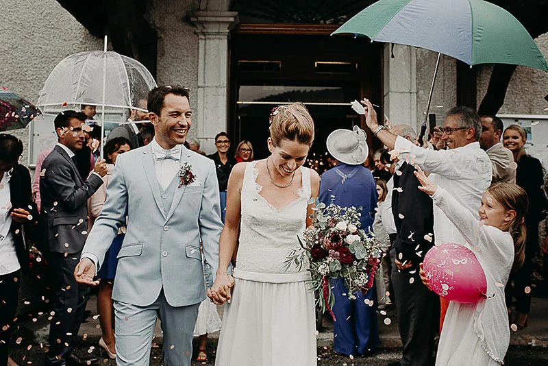 Jeunes mariés souriants sous une pluie de confettis, entourés d'invités et de parapluies devant une mairie.