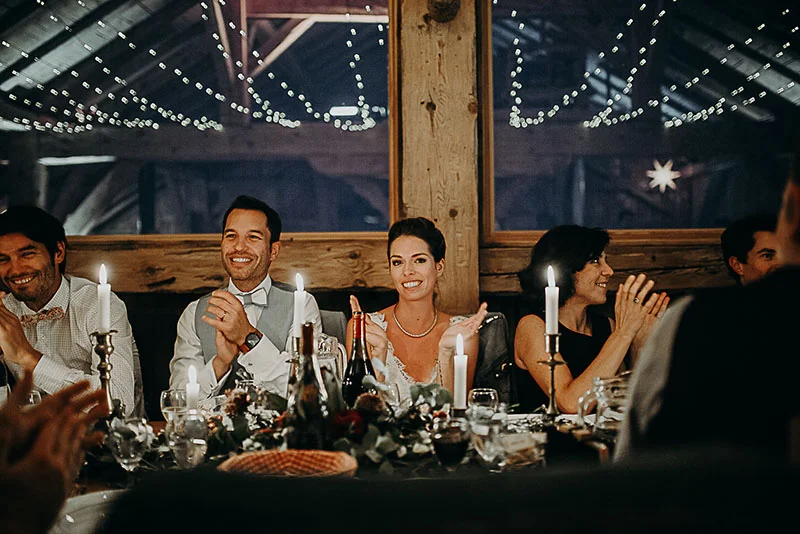 Invités souriants et élégants lors d'un dîner de mariage chaleureux sous des guirlandes lumineuses dans une grange.