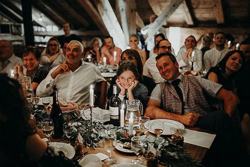 Invités souriants lors d'un dîner de mariage chaleureux dans une grange rustique aux tons boisés.