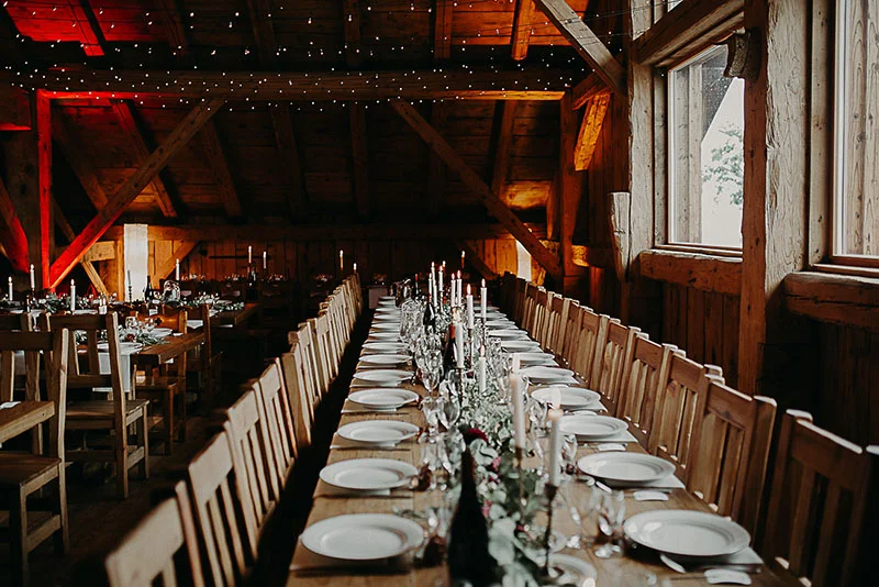 Longue table dressée dans un chalet en bois chaleureux avec bougies et lumières tamisées pour un dîner de mariage.