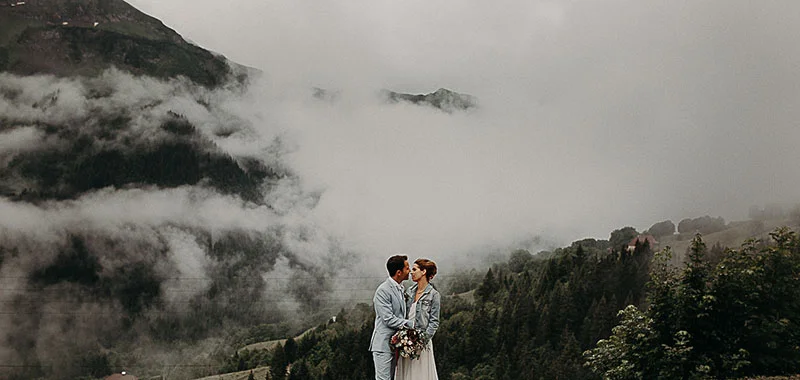 Jeunes mariés s'embrassant dans un paysage montagneux brumeux, ambiance romantique et éthérée aux tons neutres.