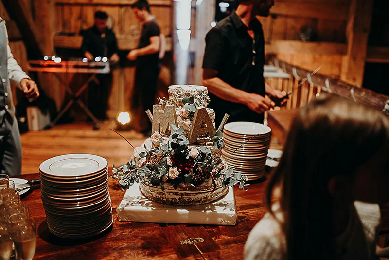 Gâteau de mariage à étages décoré de fleurs et lettres dorées sur une table en bois dans une ambiance chaleureuse.
