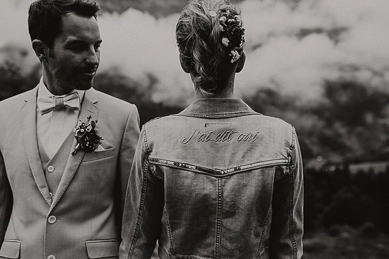 Couple de mariés en noir et blanc, la mariée de dos portant une veste en jean brodée J'ai dit oui.