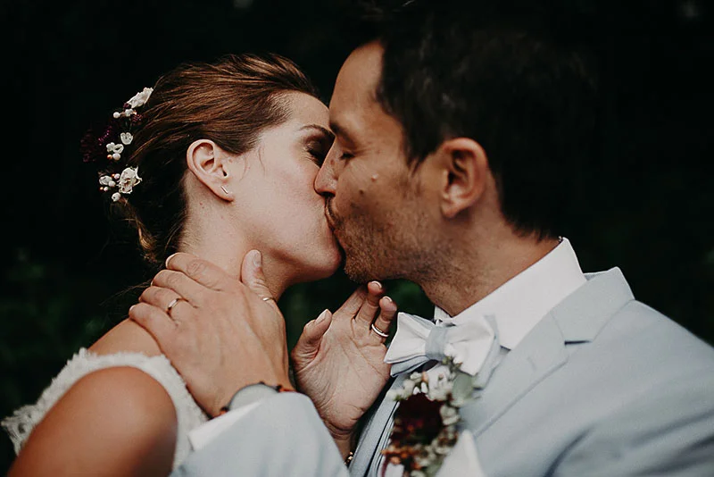 Un couple de mariés s'embrasse tendrement, elle en robe blanche et lui en costume gris clair, dans une ambiance romantique.