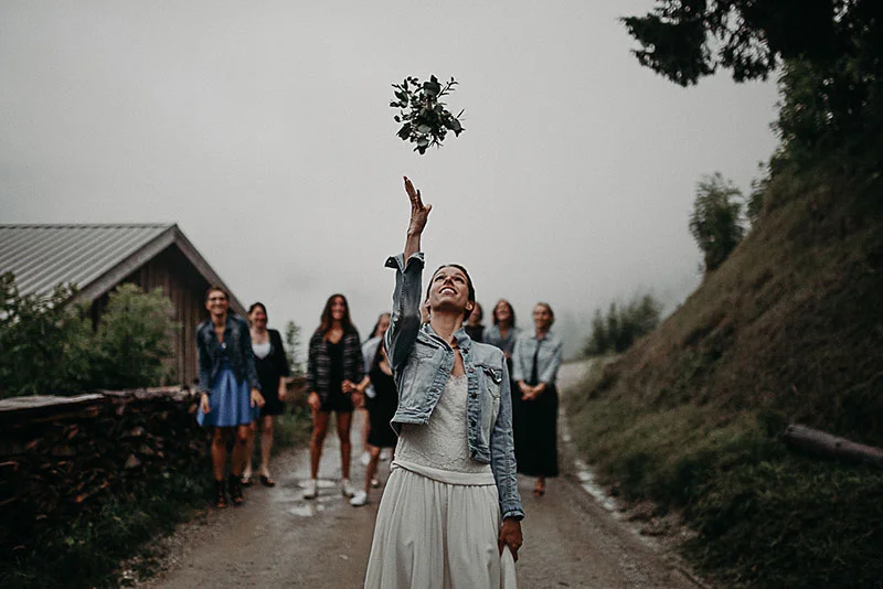 Une mariée en veste en jean lance son bouquet vers un groupe d'invitées sur un chemin rural sous un ciel brumeux.