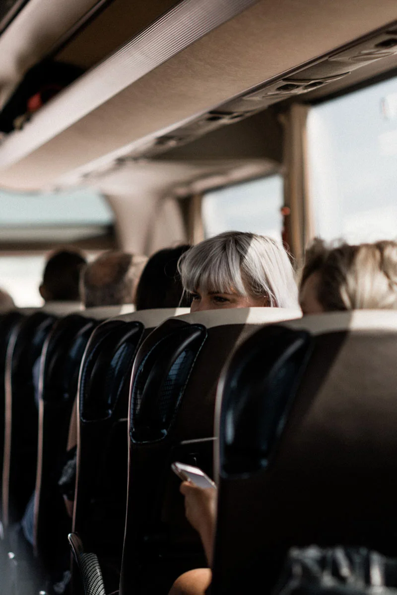 Passagers installés dans un bus aux tons neutres, avec une femme blonde regardant discrètement par-dessus son siège.