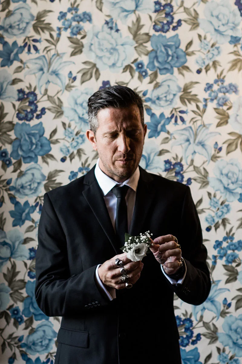 Un homme en costume noir ajuste sa boutonnière blanche devant un papier peint floral bleu et blanc.