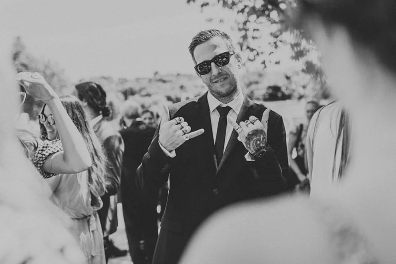 Homme souriant en costume sombre et lunettes de soleil, faisant un signe de la main lors d'un mariage en noir et blanc.