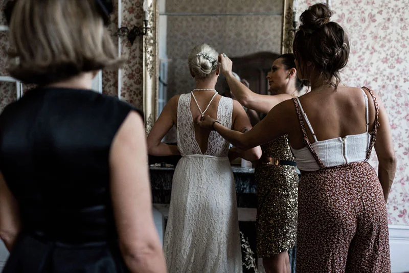 Une mariée en robe blanche se fait aider pour ses finitions par deux femmes dans une chambre à l'ambiance vintage.