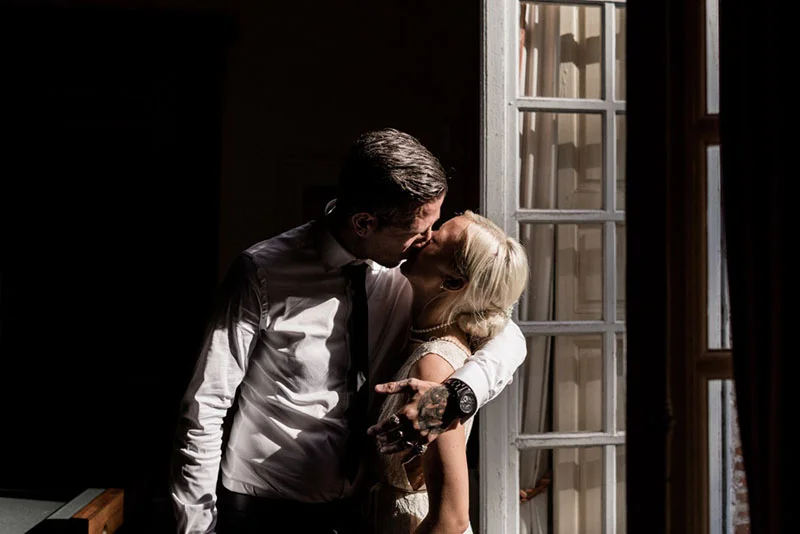 Couple s'embrassant avec passion dans un jeu d'ombre et de lumière près d'une fenêtre, ambiance romantique et intime.