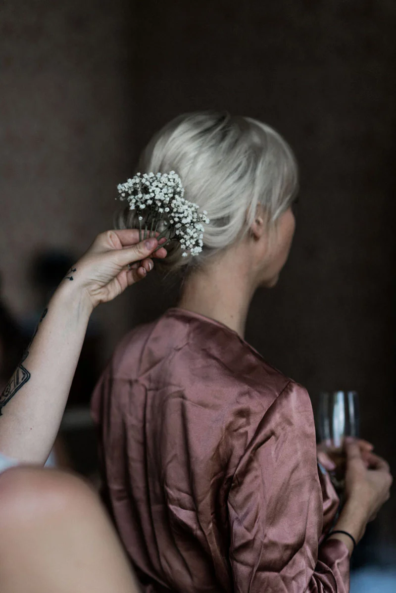 Gros plan sur une mariée en robe de chambre satinée rose poudré, dont le chignon est orné de gypsophile blanc.