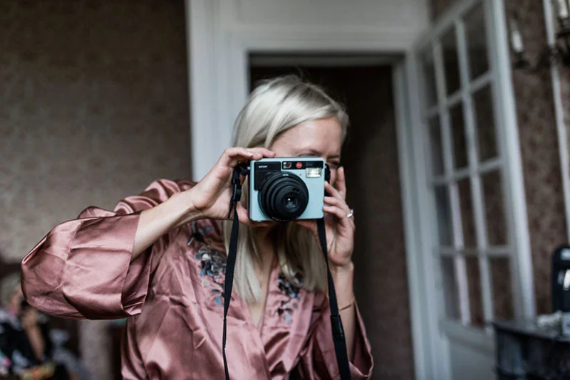 Une femme en robe de chambre rose satinée prend une photo avec un appareil argentique.