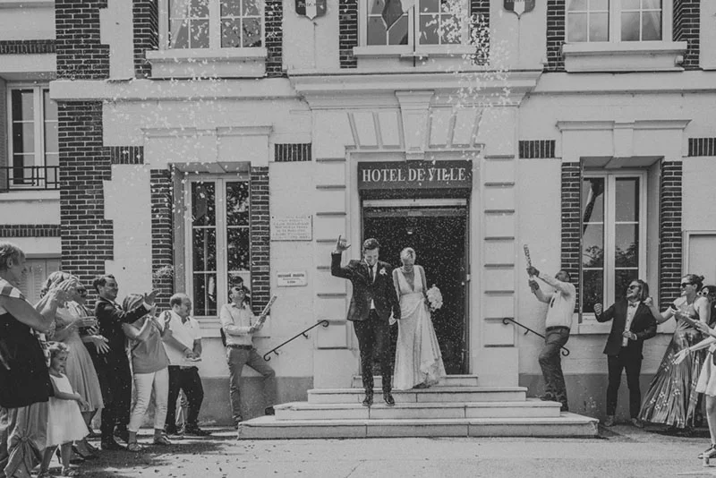 Jeunes mariés sortant d'une mairie sous une pluie de confettis, entourés d'invités, en noir et blanc.