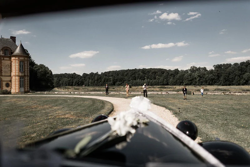 Vue depuis une voiture décorée de fleurs, des invités marchent vers un château sous un ciel bleu et lumineux.