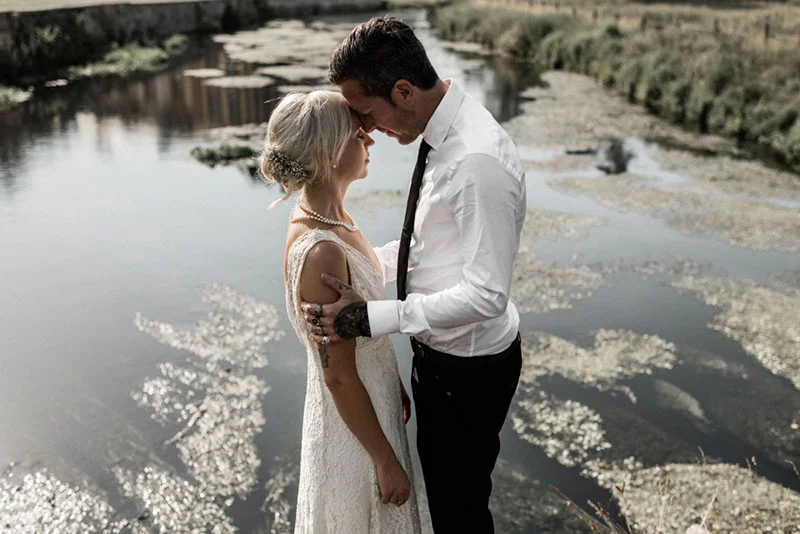 Couple de mariés s'embrassant tendrement au bord d'un étang, ambiance romantique et naturelle aux tons sobres.