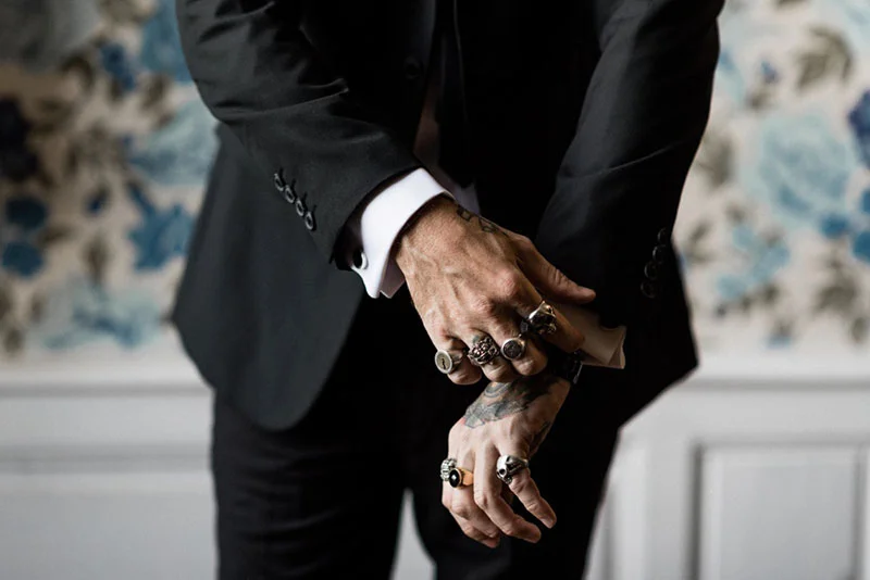 Gros plan sur les mains tatouées et bagues d'un homme en costume noir devant un papier peint floral bleu.
