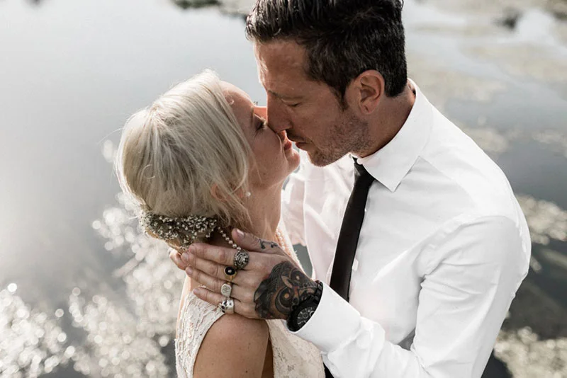 Un couple marié s'embrasse tendrement au bord de l'eau, elle en robe blanche et lui en chemise blanche et cravate noire.