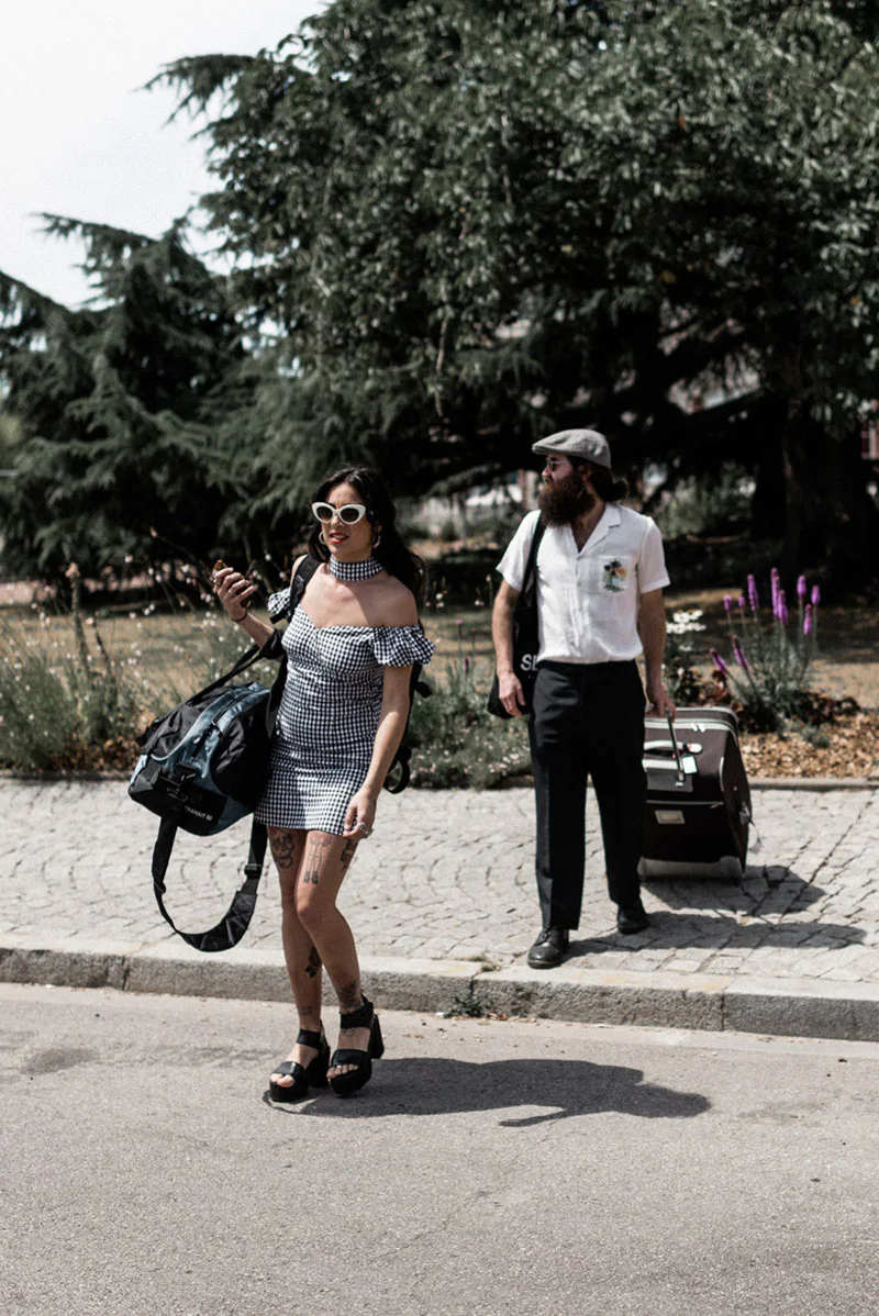 Couple stylé avec bagages marchant en extérieur, elle en robe à carreaux et lui en chemise blanche et casquette.