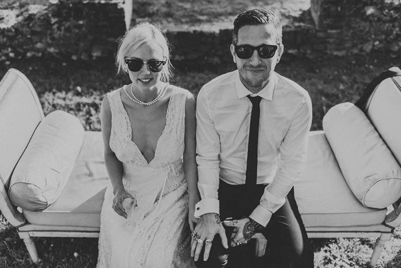 Couple marié stylé en noir et blanc, portant des lunettes de soleil, assis ensemble sur un canapé d'extérieur.