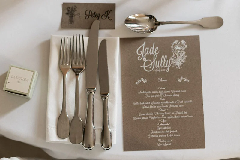 Dressage de table élégant avec menu kraft, couverts argentés et boîte Ladurée pour le mariage de Jade et Sully.