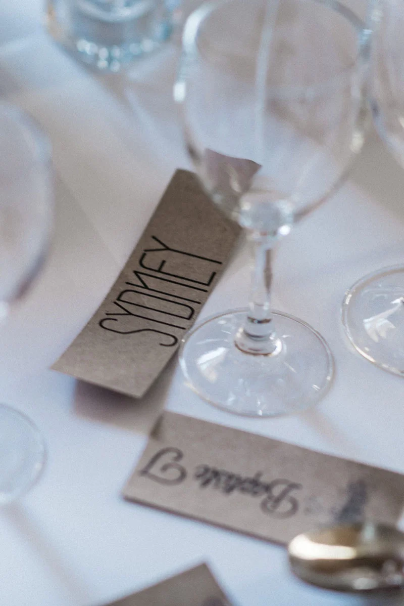 Marque-places minimalistes en papier kraft avec calligraphie noire sur une table de mariage blanche et épurée.