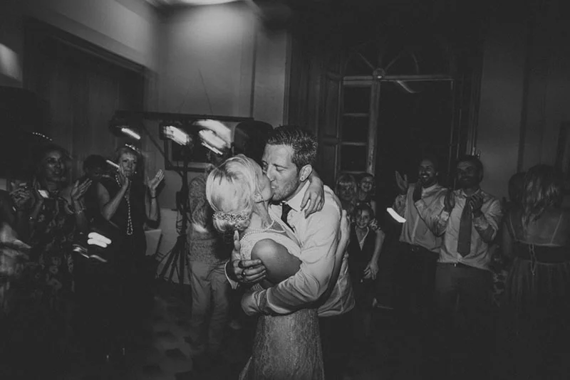 Couple de mariés s'embrassant passionnément sur la piste de danse, entourés d'invités, en noir et blanc.