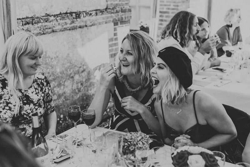 Trois femmes rient ensemble lors d'un dîner de mariage, photo en noir et blanc à l'ambiance festive et conviviale.