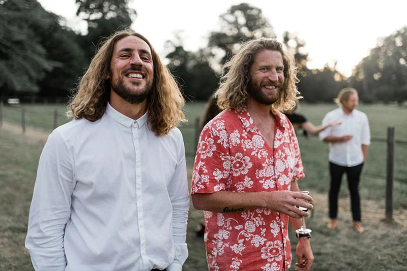 Deux hommes souriants aux cheveux longs, l'un en chemise blanche et l'autre en chemise rouge fleurie, lors d'un mariage.