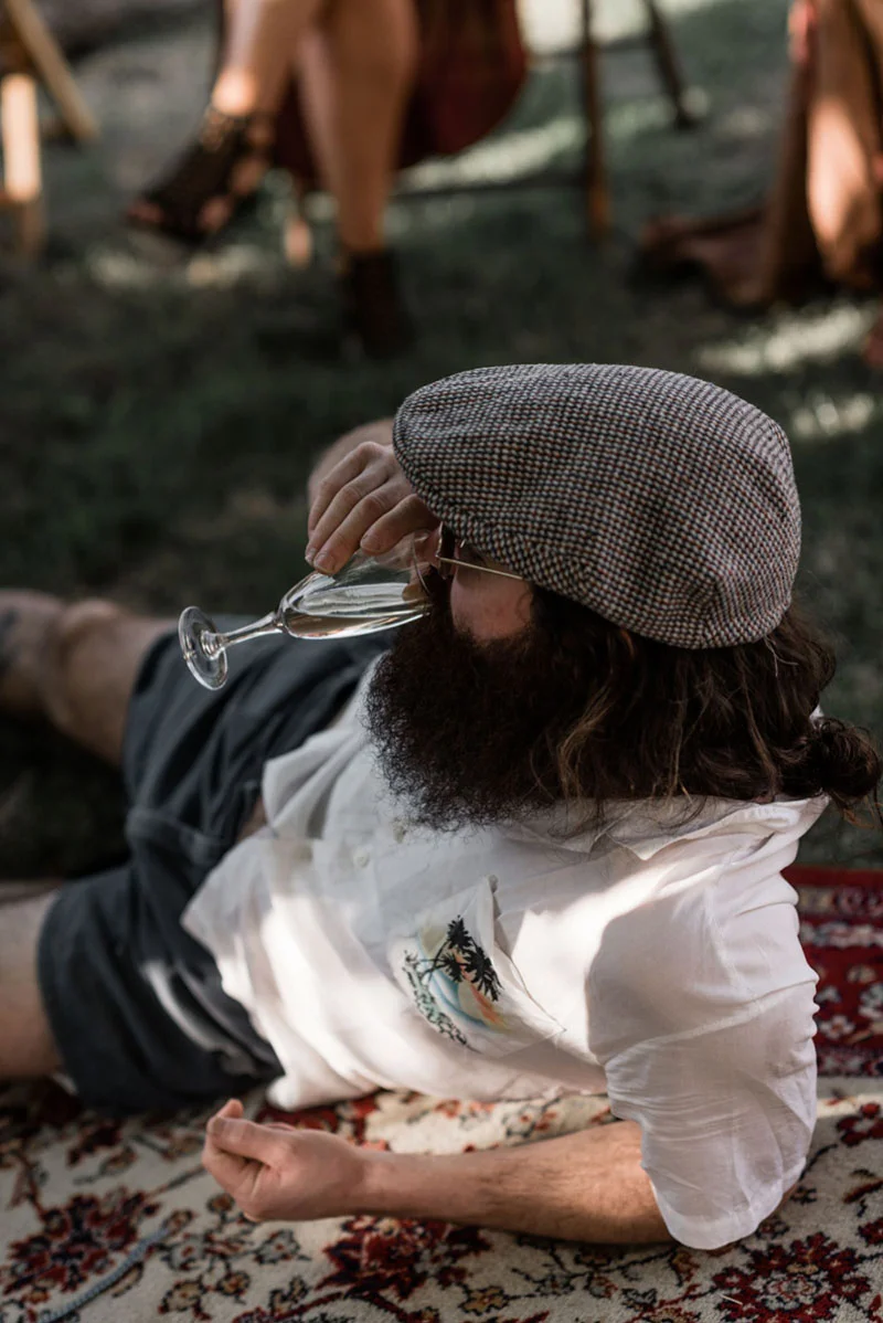 Homme barbu à la casquette buvant du champagne, allongé sur un tapis lors d'un mariage bohème en extérieur.